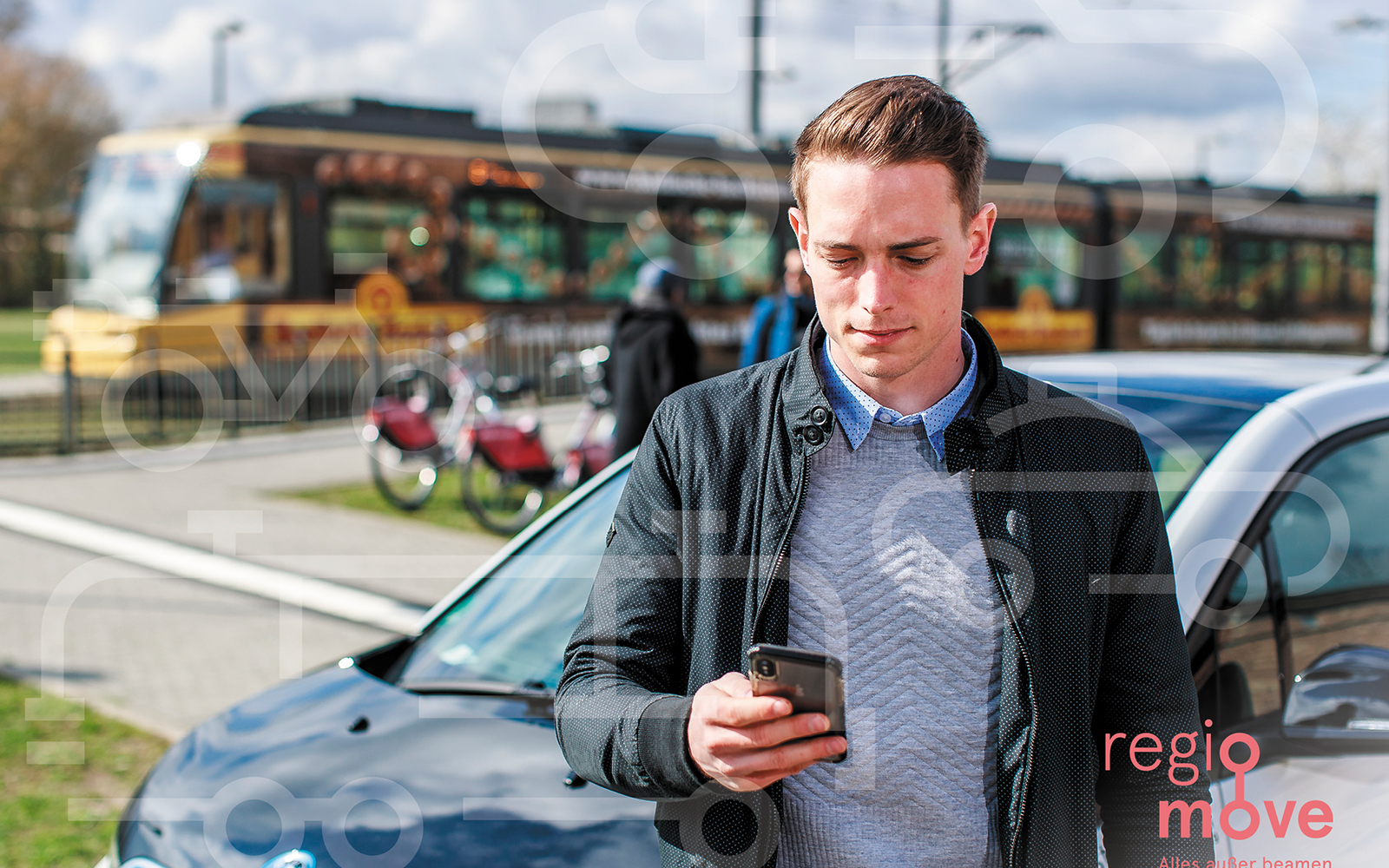 Junger Mann mit Mobiltelefon in der Hand, im Hintergrund PKW, Räder und Straßenbahn in Haltestelle