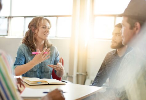 Frau mit Stift in der Hand und Notizbuch erläutert am Tisch 3 Personen Sachverhalt