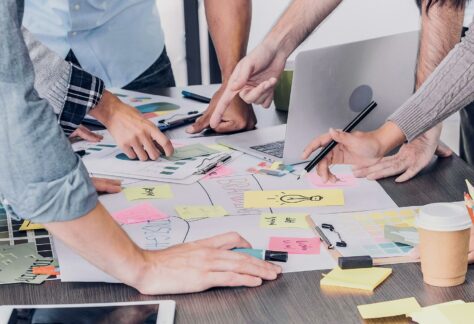 Gruppenarbeitssituation: Arbeitstisch mit Plakat und Post-Its und Händen von 5 Personen mit Laptop und Tablet