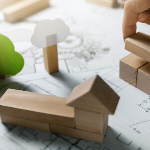 Hand stapelt Holzbauklötzchen in einem Wohn-Szenario mit Holzklätzchen, die eine Kirche bilden, Bäumen aus Holzklötzchen und Papierlaub, alles auf einem Architekturplan stehend